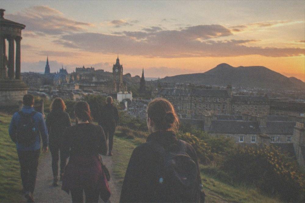 Panoramic view from a grassy hilltop across Edinburgh rooftops toward the volcanic silhouette of Arthur's Seat