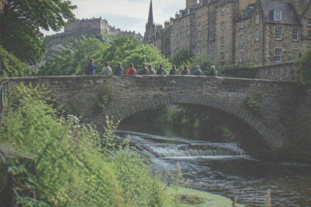 A stone bridge arching over a river with weirs, surrounded by trees and old stone buildings along the bank