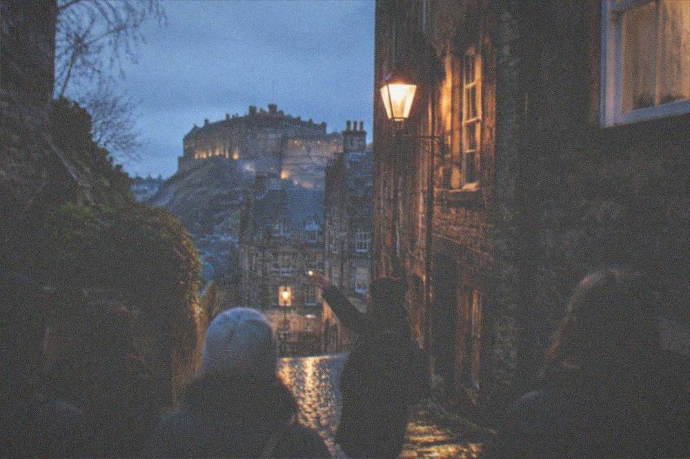 An Edinburgh close at dusk with lamplight on wet cobblestones and old tenement walls rising on both sides