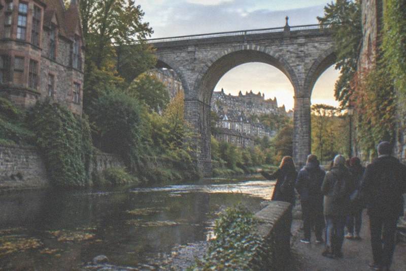 Casitas de piedra y el arroyo Water of Leith bajo los arcos de un puente de Dean Village, con hiedra trepando por los muros antiguos y un paseante junto al agua en la luz del atardecer.