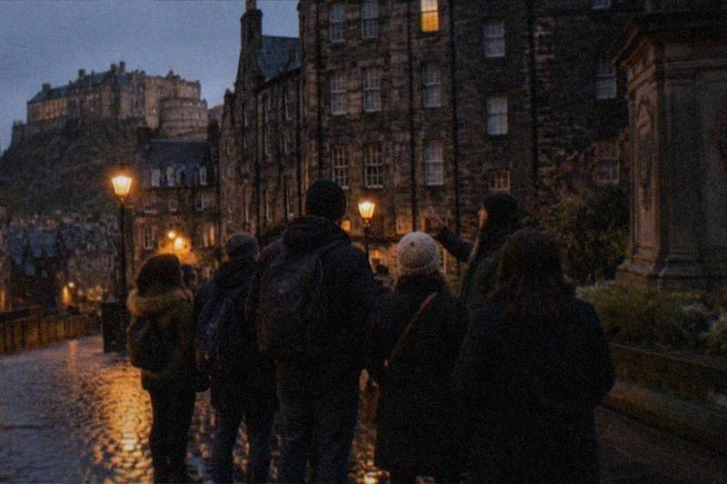 Un callejón estrecho del Old Town de noche, con los adoquines mojados reflejando la luz ámbar de las farolas bajo altos edificios ennegrecidos y una ventana iluminada en lo alto del muro de piedra.