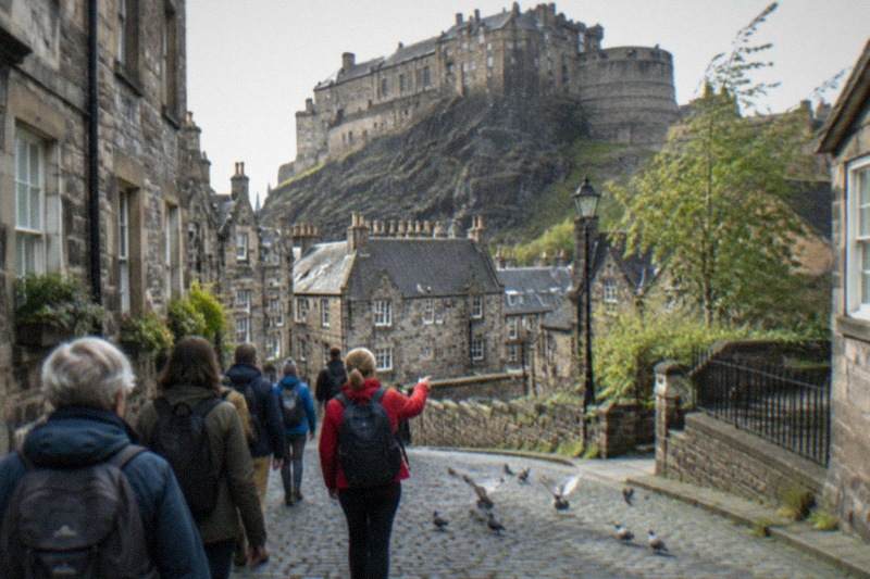 Murallas de piedra desgastada del castillo de Edimburgo, elevándose sobre la oscura roca basáltica, con nubes bajas cruzando el cielo sobre la explanada vacía y tejados descendiendo a sus pies.